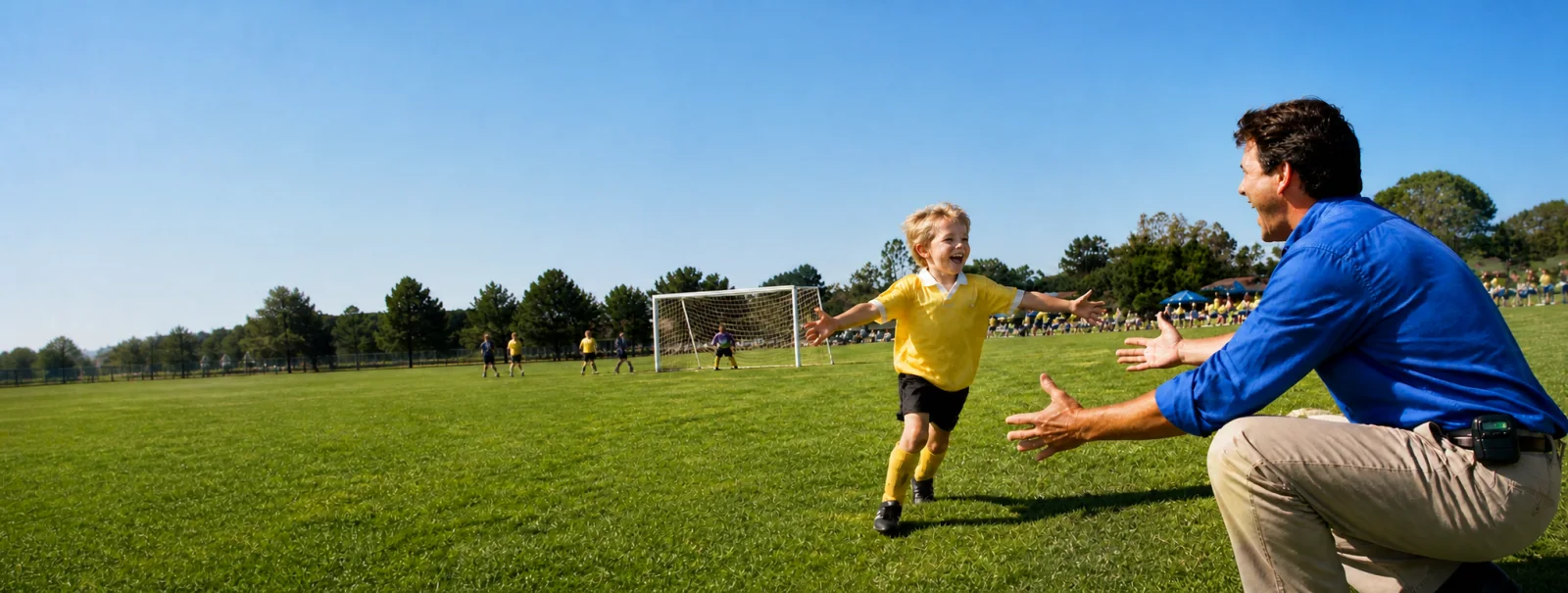 A father at the sideline opening his arms as his young son runs toward him after scoring, while the work phone at his hip shows an incoming call